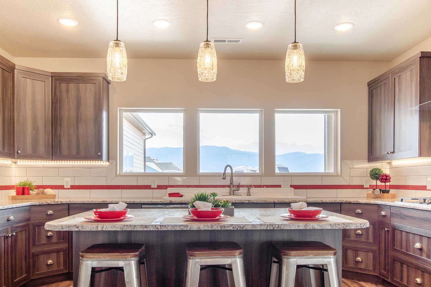 kitchen area showing tile backspace with red accents through the middle, three windows above the sink, drop ceiling lighting, three bar stools made of silver metal and wood tops