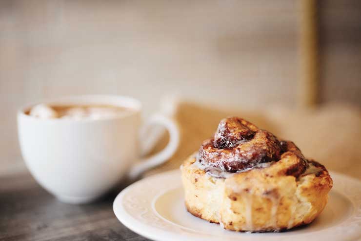 Cinnamon roll with melted icing beside a white cup with coffee and frothy milk